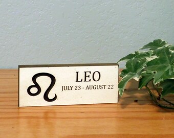 May include: A wooden sign with the Leo zodiac symbol and text. The sign is rectangular with a light background and dark brown text. The text reads "LEO JULY 23 - AUGUST 22". A green plant is in the background.