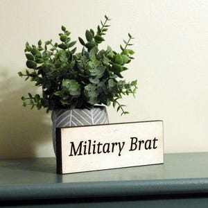 May include: A wooden sign with the text "Military Brat" in black lettering on a white background. The sign is sitting on a green surface in front of a potted plant.