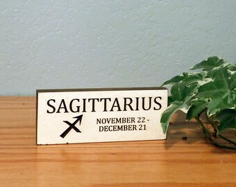 May include: A rectangular wooden sign with the word "SAGITTARIUS" in black capital letters, along with the Sagittarius symbol and the dates November 22 - December 21. The sign is on a wooden surface with a plant in the background.