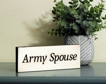 May include: White wooden sign with black text that reads "Army Spouse". The sign is sitting on a teal surface with a potted plant in the background.