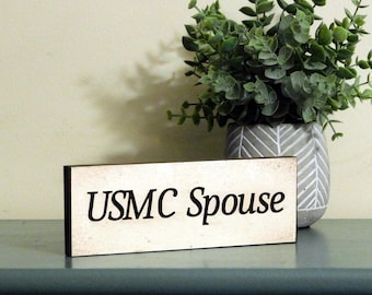 May include: A white wooden sign with black lettering that reads "USMC Spouse". The sign is sitting on a light blue surface with a green plant in a white pot behind it.