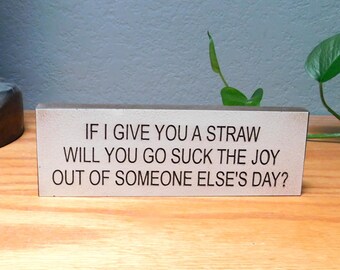 May include: A rectangular wooden sign with the text "IF I GIVE YOU A STRAW WILL YOU GO SUCK THE JOY OUT OF SOMEONE ELSE'S DAY?" in black letters. The sign is on a wooden surface.