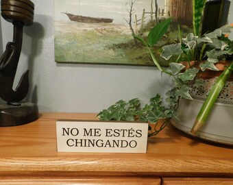 May include: A wooden sign with the text "NO ME ESTÉS CHINGANDO" sits on a wooden surface. The sign is in front of a potted plant and a painting of a boat on the water. A decorative hook is on the left.