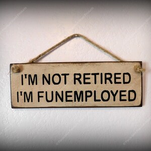 May include: A rustic wooden sign with a distressed white background and black text that reads "I'M NOT RETIRED I'M FUNEMPLOYED". The sign is hanging from a piece of twine.