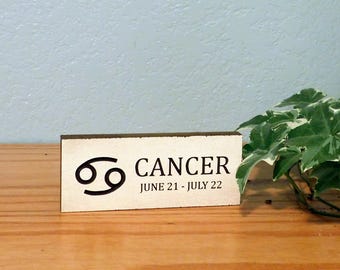 May include: A rectangular wooden sign with the Cancer zodiac symbol and text. The sign displays the Cancer symbol, the word "CANCER," and the dates "JUNE 21 - JULY 22" in black lettering. The sign is on a wooden surface.