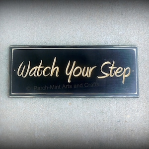 May include: Black wooden sign with white trim, featuring the text "Watch Your Step" in gold lettering. The sign is made by Parch-Mint Arts and Crafts.