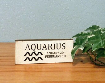 May include: A rectangular wooden sign with the word "AQUARIUS" in black, along with the dates "JANUARY 20 - FEBRUARY 18." Below the word is a black wavy symbol. The sign is on a wooden surface with green and white leafy plants in the background.