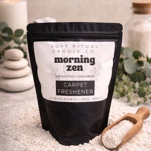 Op de afbeelding: Zwarte zak "Morning Zen" tapijtverfrisser met "Eucalyptus Spearmint" geur. De zak ligt op een oppervlak bedekt met de verfrisser. Een houten schep en een brandende kaars staan op de achtergrond. De zak bevat 850g product.