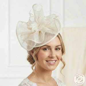 May include: A cream-colored fascinator with a layered design and feather accents. The headpiece features a wide brim and a net overlay. The woman in the image is wearing the fascinator. The fascinator is from WeddingDay Fascinators.