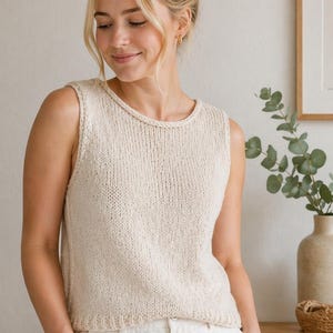 May include: Cream-colored knitted tank top with a round neckline. The sleeveless top is paired with white pants. The image is well-lit, showcasing the texture of the knitwear. The overall aesthetic is casual and comfortable.