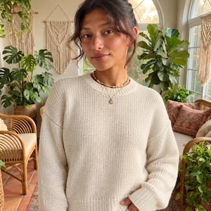 May include: A cream-colored long-sleeve sweater with a crew neckline, ribbed cuffs, and a ribbed hem. The sweater features a classic knit texture and a relaxed fit. It is paired with light-colored trousers that have a drawstring waist detail.