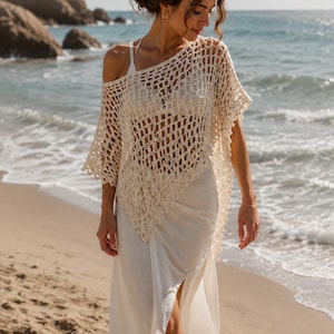 Op de afbeelding: Crèmegekleurde gehaakte poncho met een open weefpatroon. De poncho wordt gedragen over een witte jurk, waardoor een gelaagde look ontstaat. De scène speelt zich af op een zandstrand met de oceaan op de achtergrond, onder een heldere hemel.