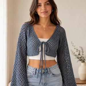 May include: A blue crochet cardigan with long, flared sleeves and a tie closure at the chest. The open-weave design of the cardigan is visible. The model is wearing a white top and blue jeans.