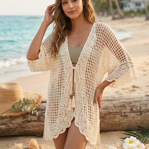May include: A cream-coloured crochet cover-up with a tie closure and tassel details. The open-weave design features a scalloped hem and three-quarter sleeves. The cover-up is shown on a beach setting.