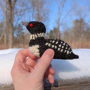 May include: A handmade, knitted loon bird figurine. The bird features a black body with white speckled markings, a black head, and a white and black striped neck. It has a gray beak and a red eye. The loon is held in a hand against a snowy background.