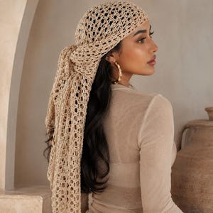 May include: A beige crochet headscarf featuring an open-knit pattern with a tied back design. The headscarf is constructed from a light-colored yarn and is styled to cover the top of the head, with long ends hanging down the back.