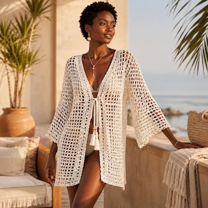 Könnte beinhalten: Cremefarbene, offene Häkeljacke mit langen, ausgestellten Ärmeln. Die Strickjacke hat einen Kordelverschluss mit Quasten und wird über einem Badeanzug getragen. Das Bild ist im Freien mit einer entspannten Strandatmosphäre aufgenommen.