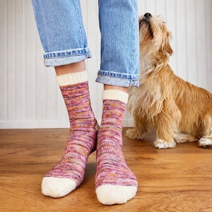 Pode incluir: Um par de meias de malha coloridas com punho e biqueira brancos, usadas com calças de ganga azuis. As meias apresentam uma mistura de fios rosa, roxos e amarelos. Um cão com pelo castanho está no fundo.