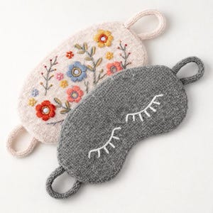 Op de afbeelding: Twee slaapmaskers. Eén is lichtroze met geborduurde bloemen in rood, geel en blauw. De andere is grijs met witte geborduurde wimpers. Beide hebben een lus om op te hangen.