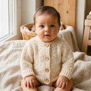 May include: A baby wearing a cream-colored, button-down knit sweater and bodysuit sits on a neutral-colored blanket. The baby is looking directly at the camera. A wooden stool and basket are in the background.