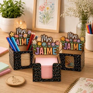 Op de afbeelding: Houten bureau-organizers met bloemaccenten en de naam "Jaime" in kleurrijke letters. De zwarte organizers met witte stippen bevatten pennen, potloden, plaknotities en paperclips. Een houten onderzetter met de tekst "3D Pixie Petal" is ook zichtbaar.