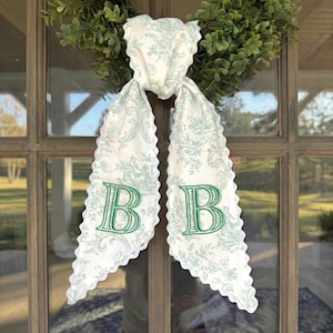 Puede incluir: Colgador de puerta de tela blanca con un estampado de toile de Jouy azul claro y bordes festoneados. La tela presenta letras mayúsculas verdes bordadas "B" a cada lado. En el fondo se ve una corona verde.
