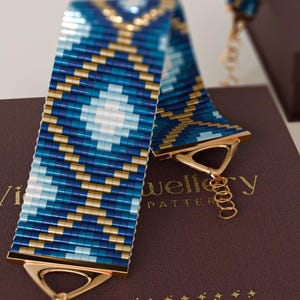 Peut inclure: Un large bracelet en perles arborant un motif géométrique dans des tons bleu foncé, bleu clair et or métallique. Le bracelet est doté d'embouts triangulaires en métal doré poli et d'une fine chaînette d'extension avec un fermoir mousqueton.