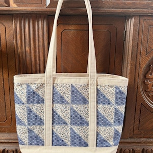 May include: A rectangular tote bag with a cream-colored base and handles. The main body features a patchwork design of blue and white floral triangles. The bag is made of fabric and has a simple, classic design.