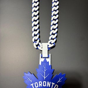 May include: A blue and white chain necklace with a large blue maple leaf pendant. The pendant features the text "TORONTO MAPLE LEAFS" in white. The chain is made of white links with blue accents.