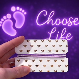 May include: A white tag with gold hearts and purple string is held in front of a purple background. The words "Choose Life" are written in neon. Two purple baby footprints are in the upper left corner.