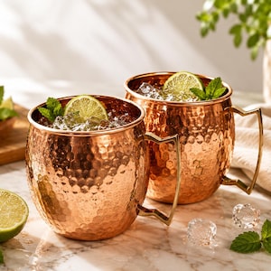 May include: Two round, hammered copper mugs with angular handles, each filled with ice, a lime slice, and a sprig of fresh mint. The mugs have a polished, reflective metallic finish and are positioned on a white marble surface with scattered ice.