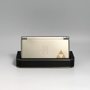 May include: A gold-colored handheld gaming console with a black and gold base. The console features a dual-screen design and a gold triangle emblem. The base has a black exterior with gold trim.