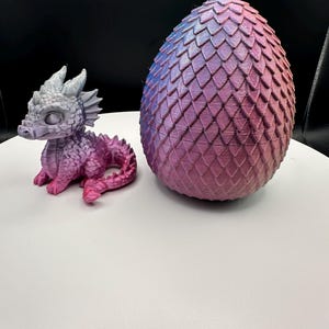 May include: A 3D-printed dragon figurine and a dragon egg. The dragon is small, with a gray head and pink body. The egg is large, with a scale pattern and a gradient of blue, pink, and purple colors. The objects are on a white surface.