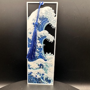 May include: A rectangular bookmark featuring a stylized depiction of a large ocean wave in shades of blue and white, with a dark background. A blue tassel hangs from the top. The bookmark is approximately 6 inches tall.