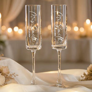 May include: Two clear champagne flutes with slender stems. Each flute is engraved with "Mrs. Smith" and the date "01.25.2026" in a graceful script. The background includes soft lighting and floral arrangements, suggesting a wedding or celebratory event.