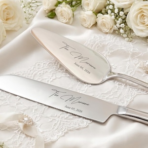 May include: A silver-toned cake knife and server set featuring a polished metallic finish. Both pieces are engraved with the text "The Mariana" and the date "June 05, 2026" in a black script and sans-serif font combination on the blades.