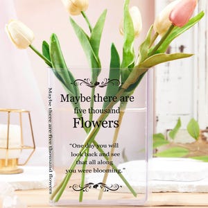 May include: A clear glass vase with several tulips in various stages of bloom. The tulips are white and pink, with green stems and leaves. The vase has text that reads "Maybe there are five thousand flowers."