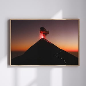 May include: A framed print of a volcano erupting at sunset. The volcano is silhouetted against a gradient sky of orange, purple, and red. The eruption emits a plume of red smoke.