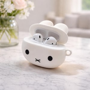 Op de afbeelding: Witte AirPods-case in cartoonstijl met een konijnenontwerp. De case heeft een ronde body met zwarte ogen en een zwarte 'x'-mond. De bovenkant gaat open en onthult witte AirPods. Een klein handvat zit aan de zijkant. Een vaas met bloemen staat op de achtergrond.