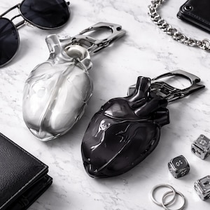 May include: Two heart-shaped keychains, one clear and one black, with silver carabiner clips. The keychains are placed on a marble surface, along with sunglasses, a wallet, and other accessories. The heart-shaped design is detailed and realistic.