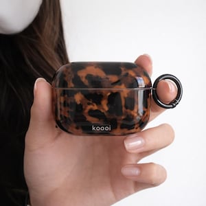 Może przedstawiać: Etui na AirPods z wzorem w żółwie skorupy z czarnym metalowym pierścieniem. Etui ma błyszczące wykończenie, a z przodu nadrukowano napis "koooi". Etui jest trzymane w dłoni osoby.