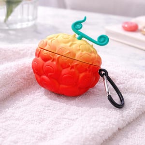 May include: A vibrant, two-toned red and yellow fruit-shaped case for wireless earbuds. It features a teal-colored stem and a black carabiner clip. The case rests on a white towel, suggesting a clean and modern aesthetic.