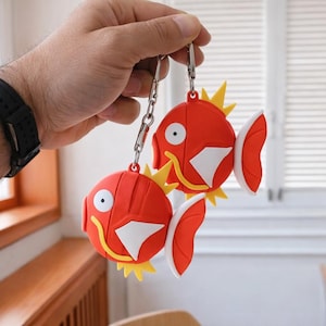 Puede incluir: Dos llaveros rojos con forma de pez con detalles blancos y amarillos. Los llaveros están sujetos a cadenas y cierres plateados. Los peces tienen un diseño de dibujos animados con ojos grandes y aletas.