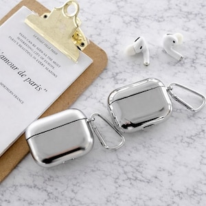 May include: Two silver metallic AirPod cases with attached carabiner clips are displayed on a marble surface. Two white AirPod earbuds are visible. A clipboard with the text "LA BELLE VILLE PARIS" and "amour de paris" is in the background.
