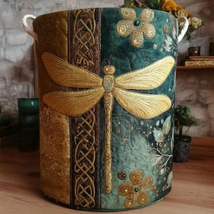 Pode incluir: Um cesto de armazenamento cilíndrico com um design acolchoado. O cesto apresenta uma grande aplicação de libélula dourada, nós celtas e enfeites florais. A paleta de cores inclui tons de verde, dourado e castanho. As alças brancas são presas no topo.