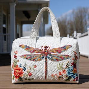 Puede incluir: Bolso tote acolchado blanco con un gran aplique de libélula colorida. El bolso presenta bordados florales y un patrón de diamantes. Tiene dos asas y un cierre de cremallera.