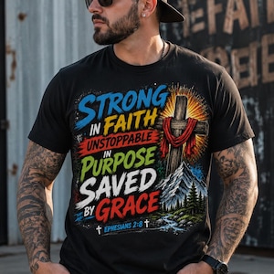 May include: A black short-sleeved t-shirt featuring a graphic print with the text STRONG IN FAITH, UNSTOPPABLE IN PURPOSE, SAVED BY GRACE, and EPHESIANS 2:8. The design includes a wooden cross with a red cloth, a mountain, trees, and a stream.