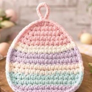 Op de afbeelding: Een pastelkleurige, gehaakte eivormige pannenlap met een lus om op te hangen. De pannenlap heeft horizontale strepen in roze, lavendel, wit en lichtblauw. De pannenlap ligt op een houten oppervlak.