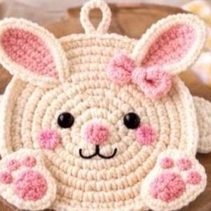 Op de afbeelding: Een crèmekleurige, gehaakte onderzetter in de vorm van een konijn met roze accenten. De onderzetter heeft een strik, binnenoren, neus, wangen en pootafdrukken in roze. Zwarte ogen en een lachende mond maken het ontwerp compleet.