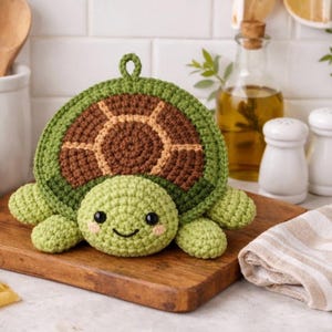 Può includere: Un sottopentola a forma di tartaruga all'uncinetto, nei toni del verde e del marrone, con un volto sorridente. La tartaruga è appoggiata su un tagliere di legno. Arredamento cucina.
